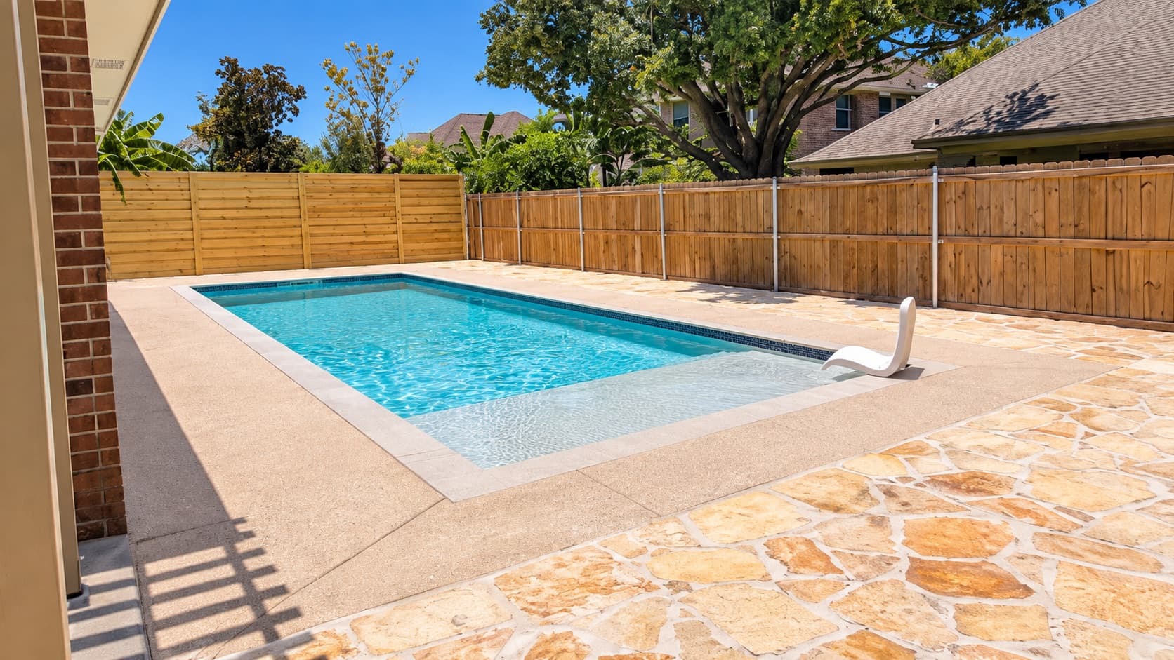 Finished backyard pool with tanning ledge, concrete deck and flagstone patio behind a brick home
